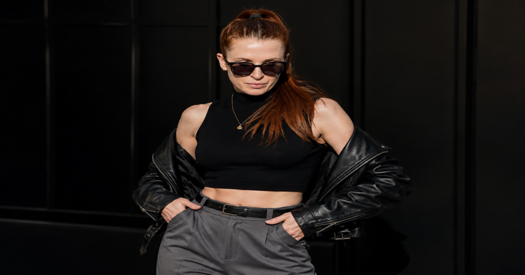 Kathleen Nimmo Lynch wearing dark sunglasses, a black sleeveless crop top, and a black leather jacket draped off her shoulders, posing confidently with hands in her pockets against a dark glass-panel background.