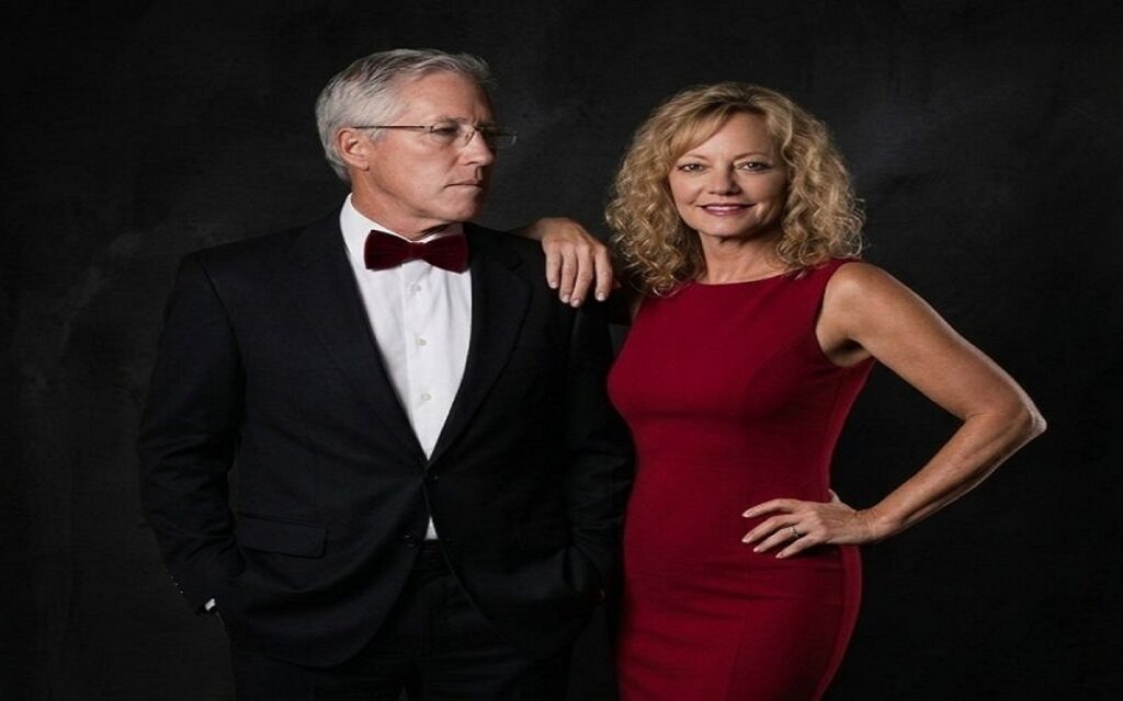 Glena Goranson and Pete Carroll posing together at a formal event, Pete wearing a black suit with a burgundy bow tie and Glena in a red dress.