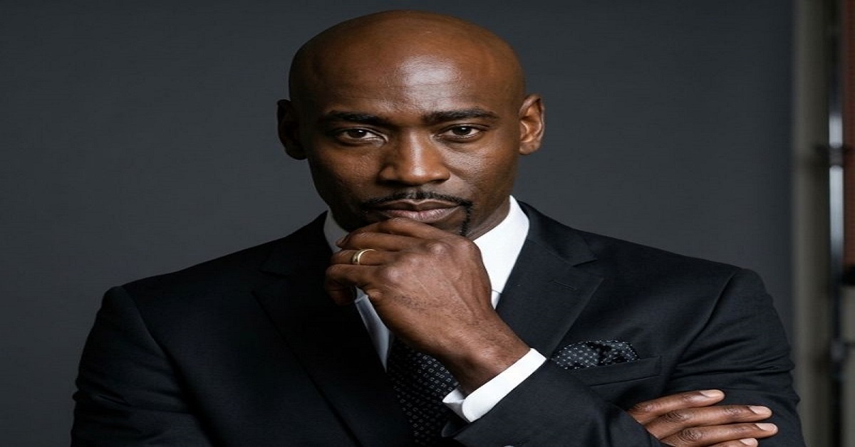 A professional headshot of Albert Ezerzer, a dedicated transportation department crew member known for his vital behind-the-scenes work on the TV show Suits (Seasons 1-3). He is a bald Black man with a neat goatee, dressed in a sharp black suit, white shirt, and patterned tie, embodying the professionalism of film industry workers.